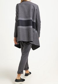 Mujer vistiendo un cardigan largo asimétrico gris oscuro con una amplia franja negra, pantalones grises a juego y zapatos negros sin cordones, caminando alejándose.