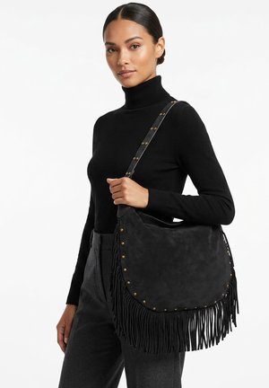 Vrouw in een zwarte coltrui en grijze broek met een zwarte suède schoudertas met franjes en messing studs.