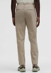 Bež tapered hlače s stražnjim džepovima, izrađene od glatkog materijala. Model nosi ležerne, teksturirane tenisice.