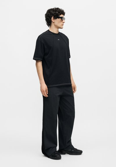 Άνδρας με σγουρά μαλλιά και γυαλιά ηλίου, φοράει μαύρο φαρδύ μπλουζάκι, φαρδιά μαύρη παντελόνια και μαύρα αθλητικά παπούτσια, στέκεται μπροστά σε λευκό φόντο.