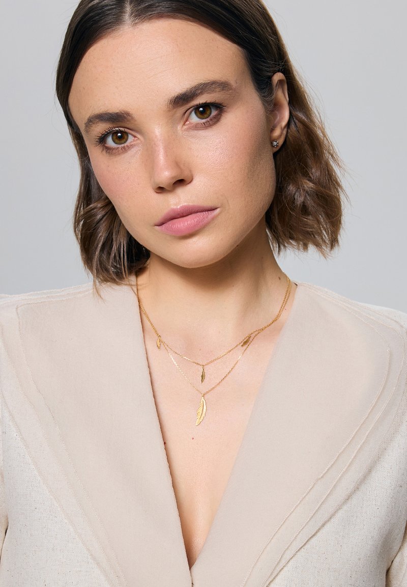 Złote naszyjniki warstwowe z pendantami w kształcie pióra, noszone na beżowej teksturowanej odzieży z głębokim dekoltem w kształcie V.