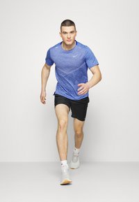 Maglietta atletica blu a maniche corte realizzata in tessuto testurizzato, abbinata a pantaloni corti neri. Modello che indossa sneakers bianche con dettagli arancioni.