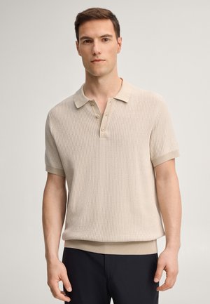 Uomo in piedi, rivolto in avanti, indossa una polo in maglia beige chiaro a maniche corte e pantaloni scuri, su sfondo chiaro e semplice.
