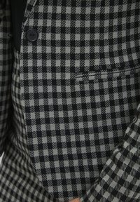 Czarny i szary w kratę żakiet z pojedynczym czarnym guzikiem, noszony na czarnej odzieży, z detalem kieszeni z przodu.