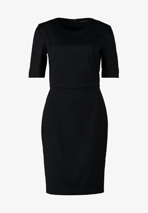 Femme en robe noire ajustée tenant une pochette beige, portant des talons noirs, debout devant un fond clair uni.