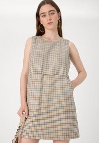 Vestuário sem mangas bege com padrão pied-de-poule em azul e castanho. Apresenta bolsos laterais e um corte descontraído. Textura de tecido suave.