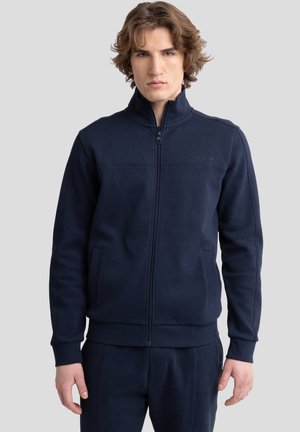 Marineblauwe ritsjack gemaakt van gestructureerde stof met een hoge kraag, zijzakken en minimale branding op de borst.