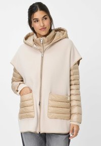 Beige hoodie met een zachte, fleece-achtige body en gewatteerde mouwen. Voorzien van twee zakken aan de voorkant en een ritssluiting.