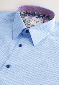 Šviesiai mėlyna tekstūruota marškinėlių tipo palaidinė su kontrastinga gėlių ir raštuota apykaklės pamušalu. Yra mėlyni mygtukai ir pritaikyta dizaino siuvimo.