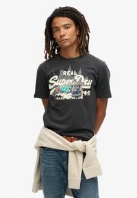 Czarny bawełniany t-shirt z graficznym nadrukiem przedstawiającym kolorowy pociąg oraz tekst. Noszony z niebieskimi dżinsami i beżowym swetrem związanym w talii.