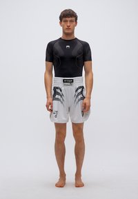 Μαύρο εφαρμοστό κοντομάνικο συμπιεστικό πουκάμισο και λευκό σορτς με μαύρα φυλετικά σχέδια. Το μοντέλο στέκεται ξυπόλυτο μπροστά σε απλό φόντο.