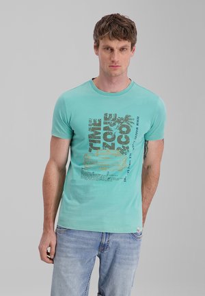 Young man with short brown hair wearing a turquoise graphic t-shirt and light blue jeans standing against a plain gray background.