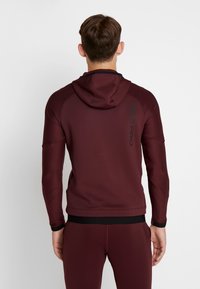 Bordeaux Nike Pro hoodie med et struktureret design, forlængede ærmer og en sort logo-detalje på ryggen, med hætte og tætsiddende manchetter.