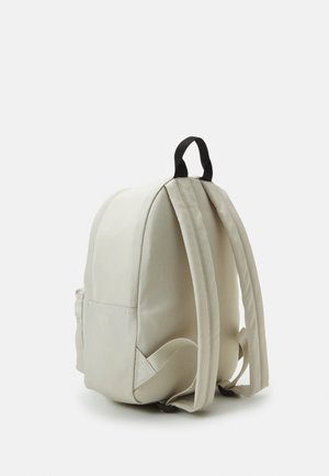 Zaino beige chiaro con spallacci imbottiti, tasca laterale e maniglia a loop in tessuto nero su sfondo bianco semplice.