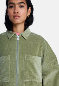 Φωτεινό πράσινο τζάκετ από κορδούρα με φερμουάρ μπροστά, γιακά με μυτερές άκρες και δύο μπροστινές τσέπες με πατ. Το ύφασμα έχει ριγωτή υφή.