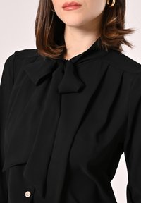 Blusa nera con un fiocco al collo, tessuto morbido, spalle plissettate e un dettaglio con un pulsante in vita.