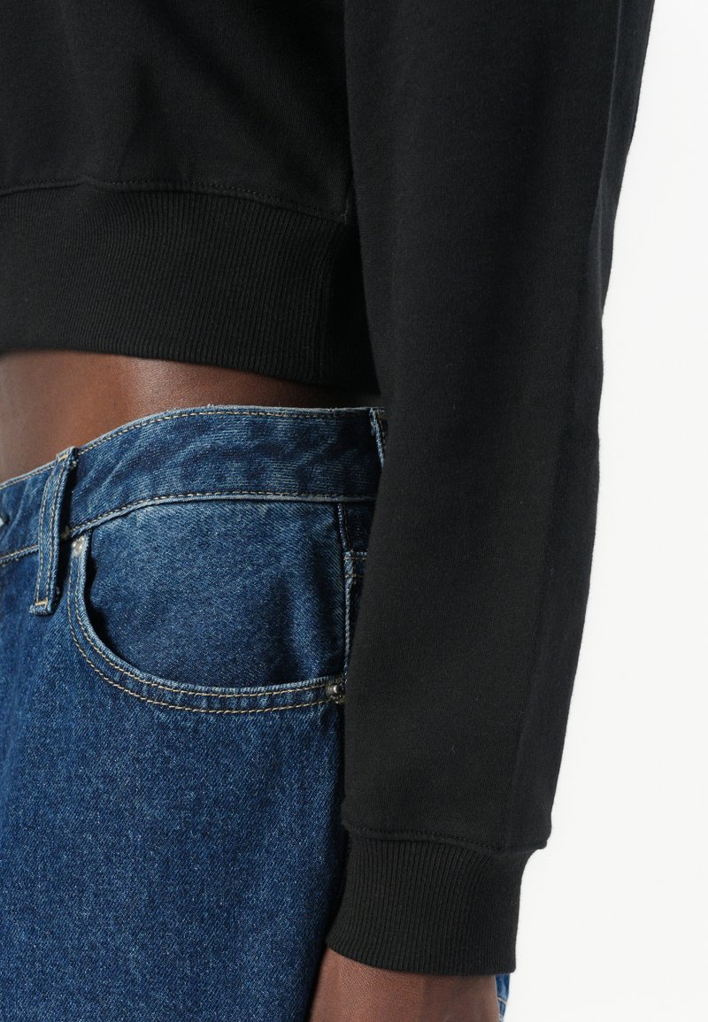 Sudadera recortada negra con puños acanalados, usada sobre jeans de denim azul claro con un acabado desgastado y costuras visibles a lo largo de los bolsillos.