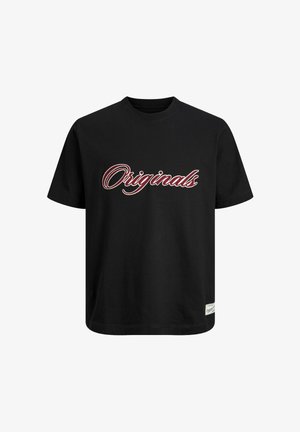 Adolescent garçon aux cheveux bouclés portant un t-shirt noir "Originals", un jean gris ample et des chaussures marron, debout devant un fond blanc uni.