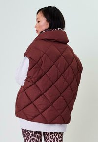 Hnedý prešívaný puffer vest s vysokým golierom a bezrukávovým dizajnom, nosený cez šedý top, skombinovaný s legínami s leopardím vzorom.