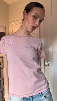 Γυναίκα με τα μαλλιά της χτενισμένα προς τα πίσω φοράει μεγάλα σκουλαρίκια κρίκους, ένα ανοιχτό ροζ μπλουζάκι με ένα μικρό σχέδιο και ανοιχτόχρωμο τζιν μέσα σε κλειστό χώρο.