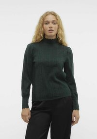 Stickad grön tröja med en hög, vågig krage, texturerade vertikala ränder och långa ärmar, tillsammans med svarta byxor.
