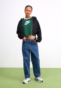 Black zip-up hoodie with grey accents, green Nike crop top, loose blue jeans, and silver-black sneakers. Plain backdrop and green flooring.