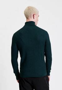 Maglione lavorato a maglia verde scuro con collettone alto, motivo testurizzato e polsini a coste. Presenta una vestibilità affusolata e dettagli sottili sull'orlo.