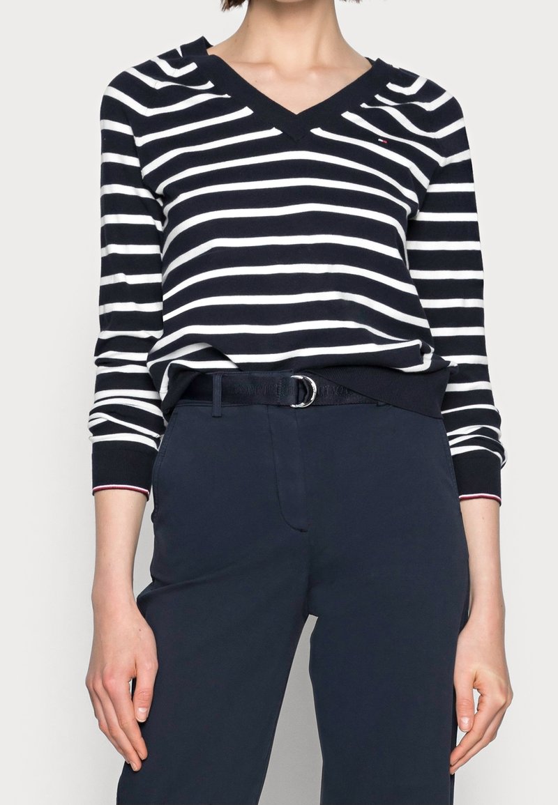 Personne portant un pull à col en V rayé bleu marine et blanc avec un pantalon bleu marine et une ceinture noire, cadrée à mi-torse.