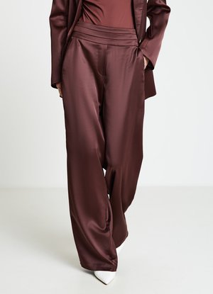 Pantalon large en satin bordeaux avec une ceinture plissée, une texture lisse et une coupe ajustée, associé à des chaussures blanches à bout pointu.