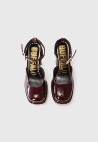 Tumed burgundia läikivast nahast plokknõelaga sandaalid, millel on pahkluu rihmad ja kuldne lukuga pannal, mis on mustadel taldadel märgistatud "BUFFALO".