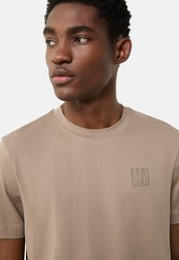 Beige bomuld t-shirt med rund hals og korte ærmer, med et diskret præget logo på brystet og glat stoftekstur.