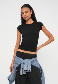 Fekete, testhezálló rövid ujjú póló, kis fémes részlettel a nyakkivágásnál. Laza fekete nadrággal és megkötött farmerdzsekivel párosítva.