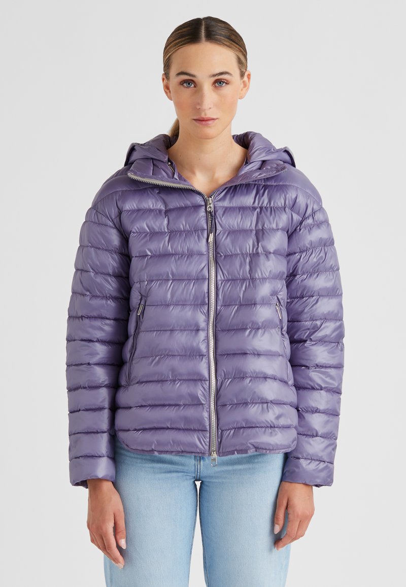 Frau in einer glänzenden, lavendelfarbenen Steppjacke mit Kapuze, zugezippt, kombiniert mit hellblauen Jeans, die vor einem schlichten Hintergrund steht.
