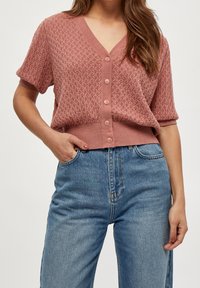 Dammig rosafärgad stickad cardigan med V-ringning, korta ärmar och fem knappar framtill, bärs över blå högmidjade jeans.