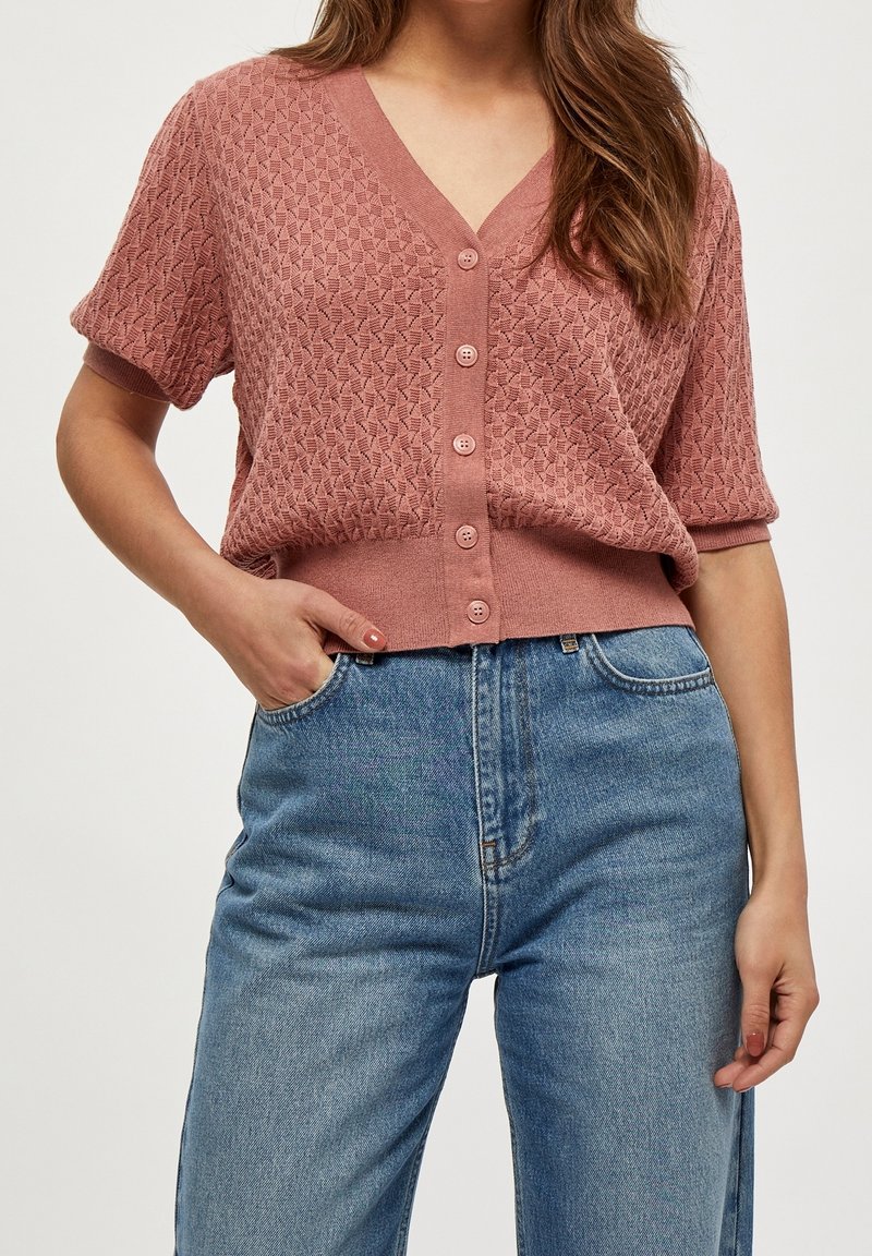 Dammig rosafärgad stickad cardigan med V-ringning, korta ärmar och fem knappar framtill, bärs över blå högmidjade jeans.