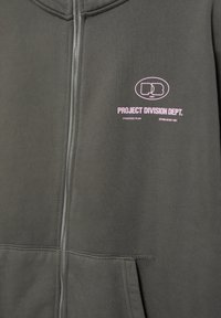 Szürke cipzáros kapucnis pulóver rózsaszín felirattal: "PROJECT DIVISION DEPT. STRATEGIC PLAN ESTABLISHED 1991" és egy kör alakú logó a felirat felett.