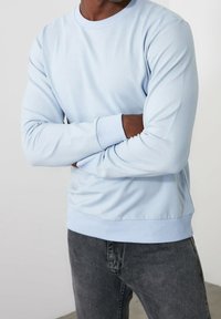 Sweat-shirt bleu clair à manches longues, col rond, poignets et ourlet côtelés. Porté avec un pantalon gris foncé, mettant en valeur une texture de tissu lisse.