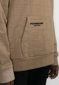 Sudadera color beige hecha de tejido suave, con un bolsillo frontal y un patrón de texto en relieve. Logo negro "PETROLIND ICE RACING" en el pecho.