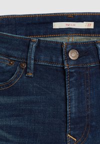 Jean en denim slim-fit de couleur bleu foncé, présentant des coutures orange, une fermeture à bouton en métal et une étiquette avec des informations sur la taille et le style.