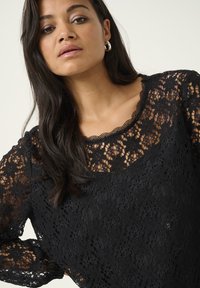 Cămașă din dantelă neagră cu un model floral, decolteu cu margine zimțată și mâneci lungi. Textură ușoară, cu tăietură lejeră.
