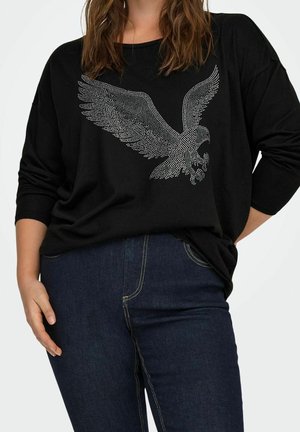 Femme portant un haut noir à manches longues avec un motif d'aigle clouté argenté et un jean bleu foncé, debout avec une main posée sur la hanche.