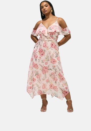 Frau in einem rosa Blumenkleid mit ausgeschnittenen Schultern und Rüschen, steht mit den Händen in der Hüfte, trägt goldene Sandaletten mit Knöchelriemen und goldene Ohrringe.