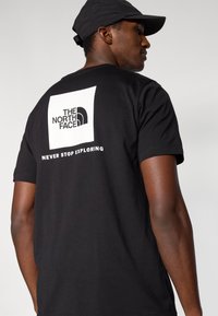 Μαύρο βαμβακερό μπλουζάκι με λευκό τετράγωνο γραφικό που φέρει το λογότυπο "THE NORTH FACE" και το κείμενο "NEVER STOP EXPLORING" στην πλάτη.