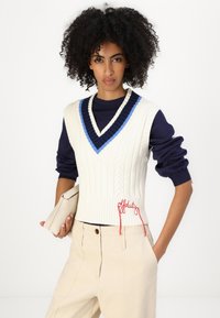 Knitted, cream v-neck vest with blue and navy accents, ribbed texture, and red "off duty" embroidery. Worn over a navy long-sleeve top.