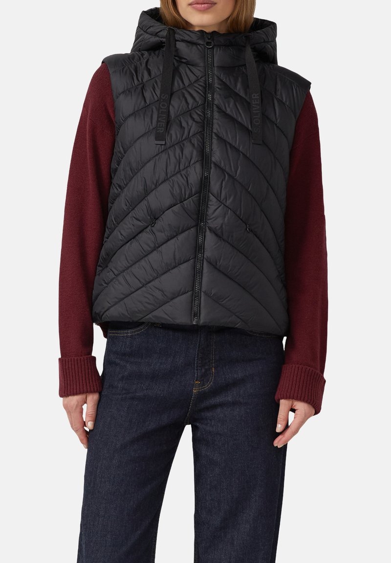 Frau in einem schwarzen, gesteppte, ärmellosen Kapuzenweste über einem langärmeligen bordeauxfarbenen Pullover und dunkelblauen Jeans vor einem weißen Hintergrund.