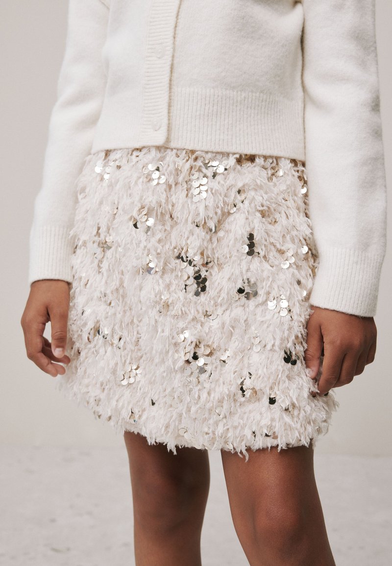Bijeli kardigan preko bež suknje sa šljokicama, izrađene od teksturiranog, mekanog tkanja s naboranim izgledom i okruglim naglascima šljokica.