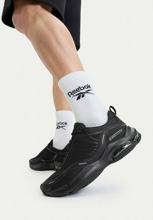 Nogi w czarnych butach Reebok z widoczną poduszką powietrzną i białych skarpetkach z logo Reebok, na jednolitym tle.