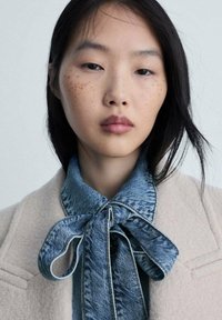 Cappotto beige con colletto a papillon in denim. Il colletto presenta un motivo testurizzato e una cucitura a contrasto, che si abbinano al morbido tessuto del cappotto.