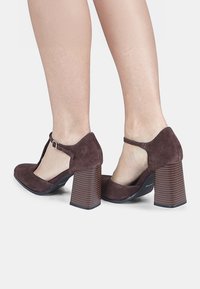 Zapatos de tacón de ante marrón con un diseño de tira en T, puntera redondeada y tacones de bloque de madera; presentan lados expuestos para un aspecto contemporáneo.