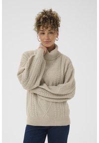Beige kaapeknitted turtleneck-swearer med brede ærmer, strukturerte mønstre og ribbestrikket kant. Kombinert med mørke jeans på en nøytral bakgrunn.